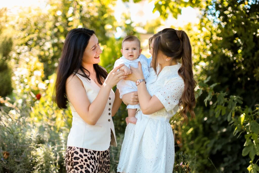 Two moms smiling at a baby in a sunny garden